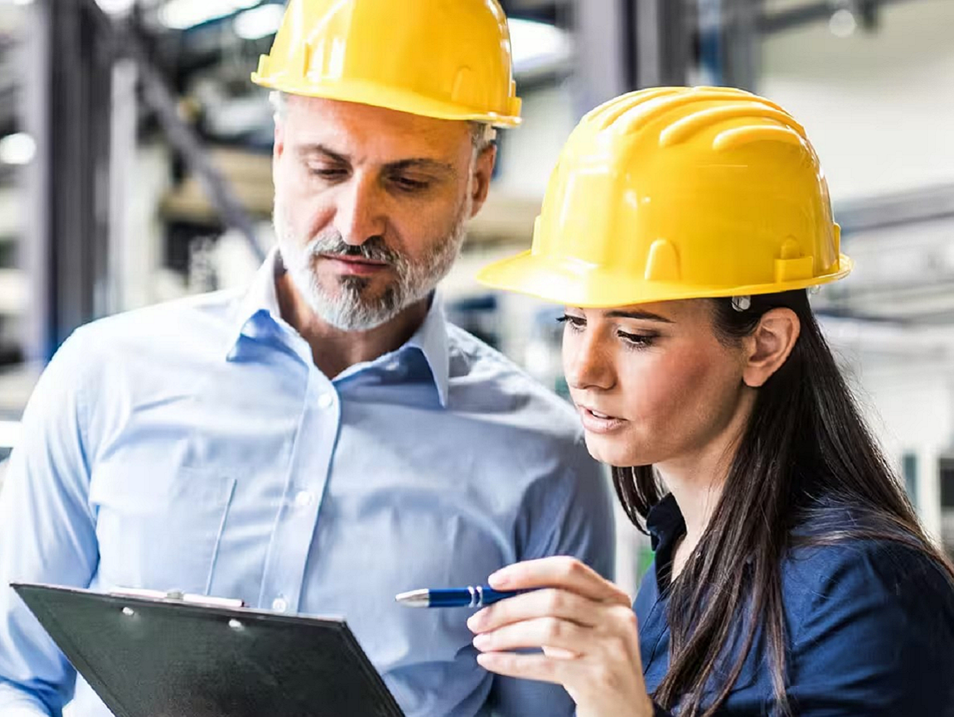 Two employees wearing safety helmets looking at a datasheet on a shop floor