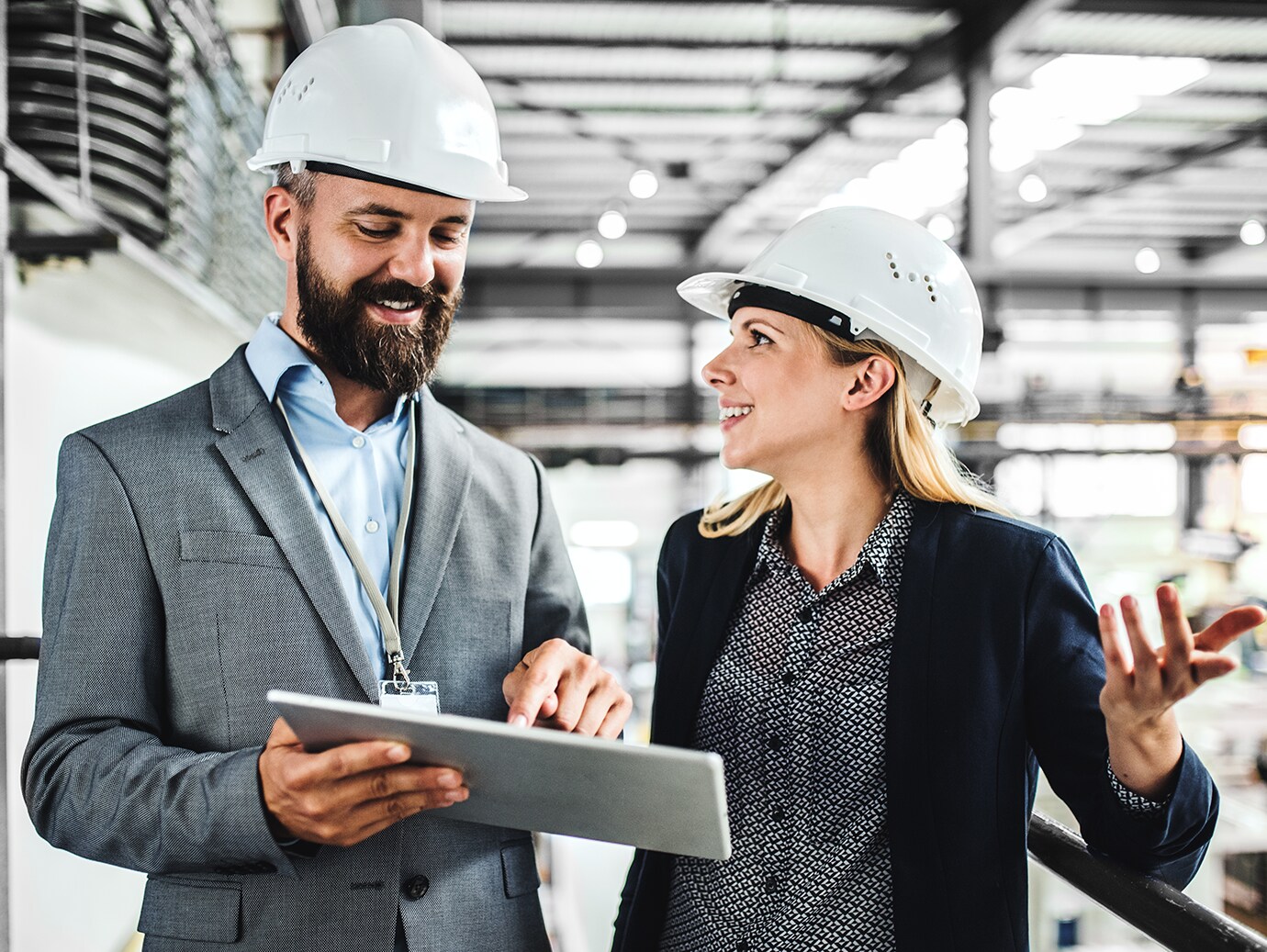 Two people discussing holding a tablet on a factory floor wearing white safety helmets